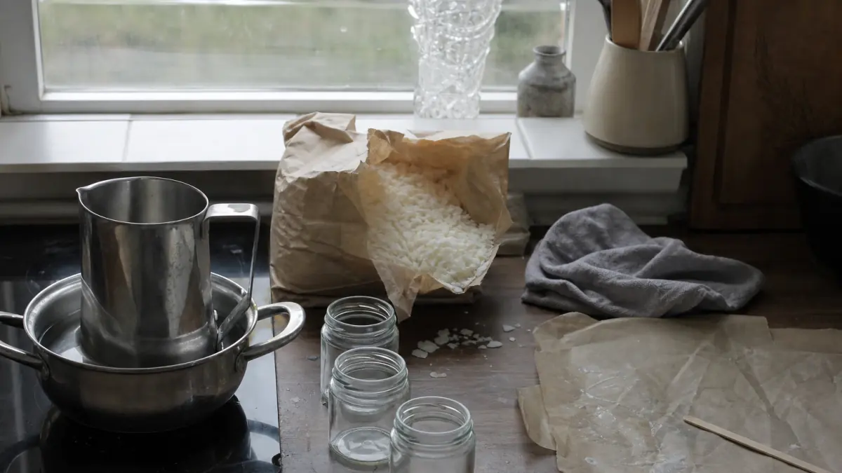 Kuchenny blat w pochmurny dzień: stalowy dzbanek w garnku z wodą na płycie, rozdarta papierowa torba płatków wosku, trzy puste słoiki, szara ścierka i papier do pieczenia z zastygłymi kroplami